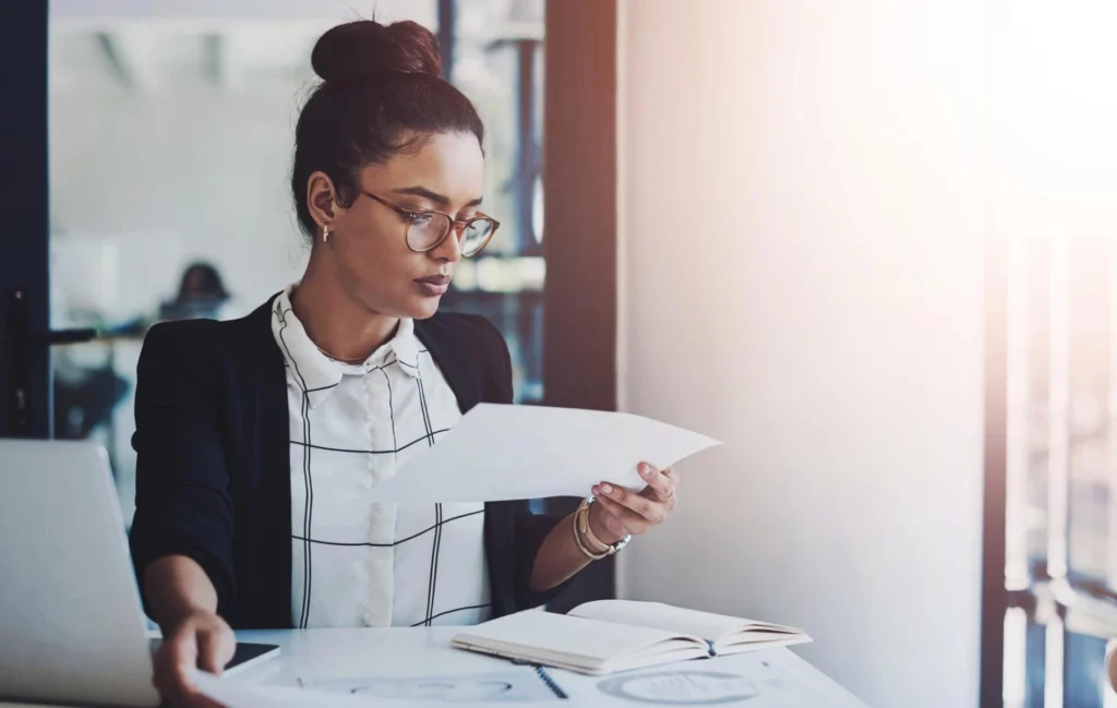 Mulher de cabelo preso, usando óculos e roupa formal, analisando documentos em ambiente de escritório, relacionado à lei orçamentária anual 2025.